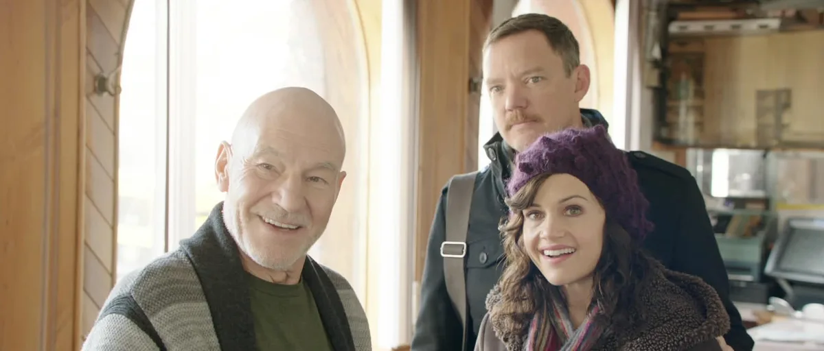 three people are in a warmly lit wooden room, with a bald man smiling broadly in the foreground, a man with a mustache…
