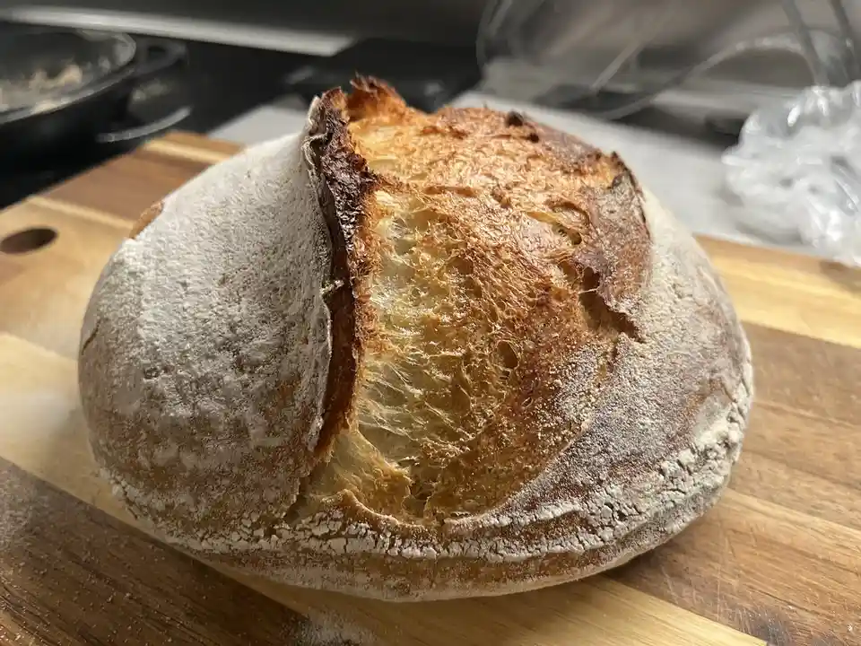 A round, rustic loaf of bread with a golden-brown, cracked crust and flour dusting sits on a wooden cutting board. The…