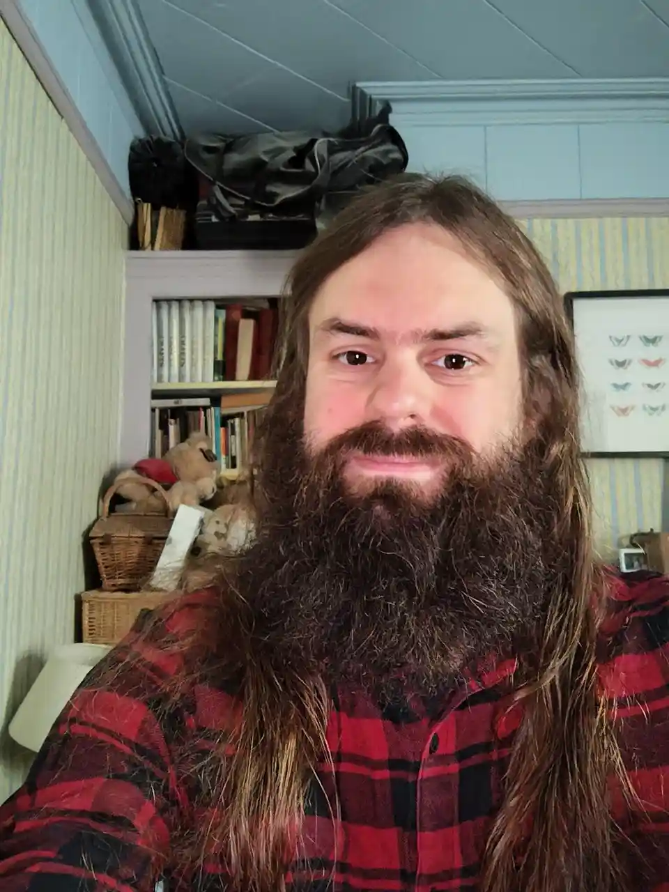 A man with long brown hair and a full beard, wearing a red and black plaid shirt, looks directly at the camera with a slight…