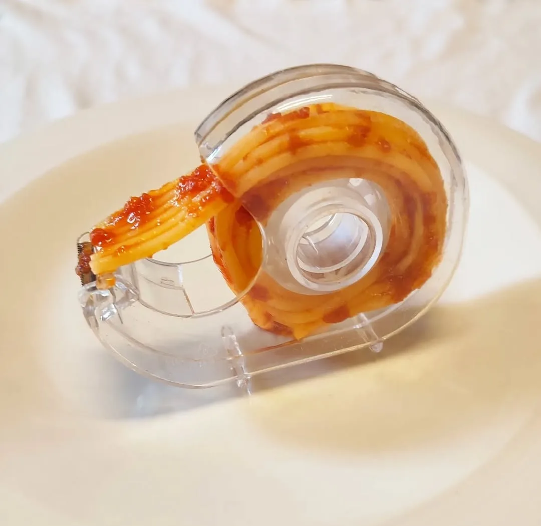 A clear tape dispenser holds a roll of cooked spaghetti noodles coated in red tomato sauce, resembling adhesive tape, placed…