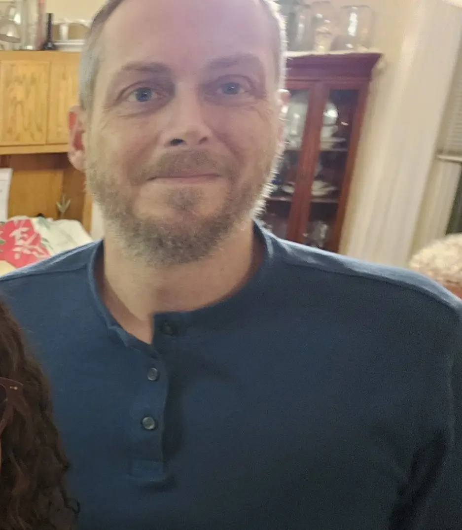 A close-up selfie of a middle-aged man with short hair and a beard, wearing a dark blue henley shirt, looking directly at…