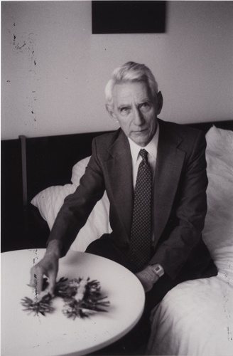 a black-and-white photo shows an older man in a suit and tie sitting on a bed, reaching toward a small, spiky, unattractive…