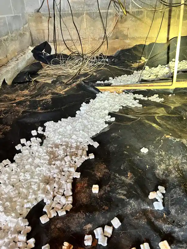 A messy basement floor covered in black plastic sheeting, tangled wires, and a large spill of white, crumpled packing…
