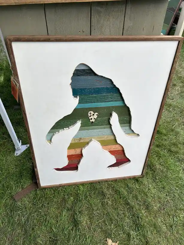 A framed wooden art piece displays a silhouette cutout of Bigfoot sitting on a colorful striped background, with a small…
