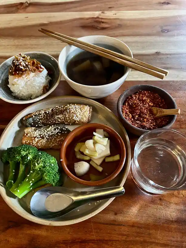 A wooden table holds a meal with a plate of grilled fish, broccoli, and a small bowl of pickled vegetables, alongside a bowl…
