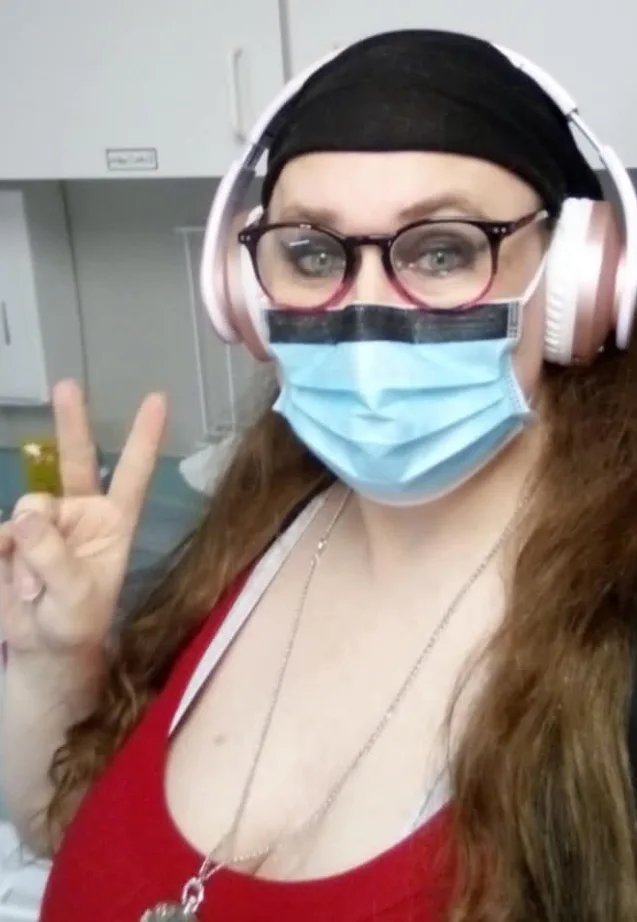 a woman with long brown hair, wearing a black headband, glasses, and large pink headphones, poses with a peace sign while…