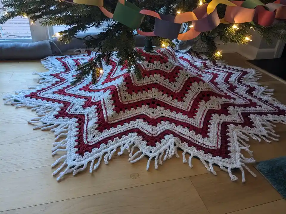 A red and white crocheted star-shaped tree skirt with fringe lies on a wooden floor beneath a decorated Christmas tree with…
