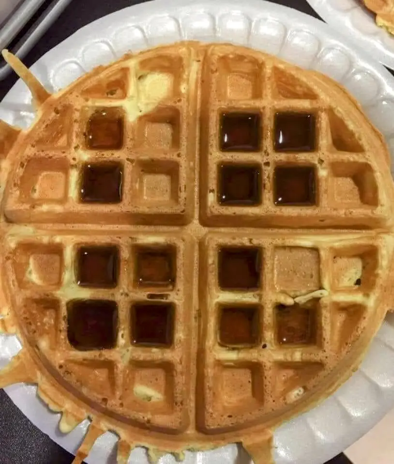 A close-up photo shows a golden-brown, round waffle on a white paper plate, with syrup pooling in its square pockets and…