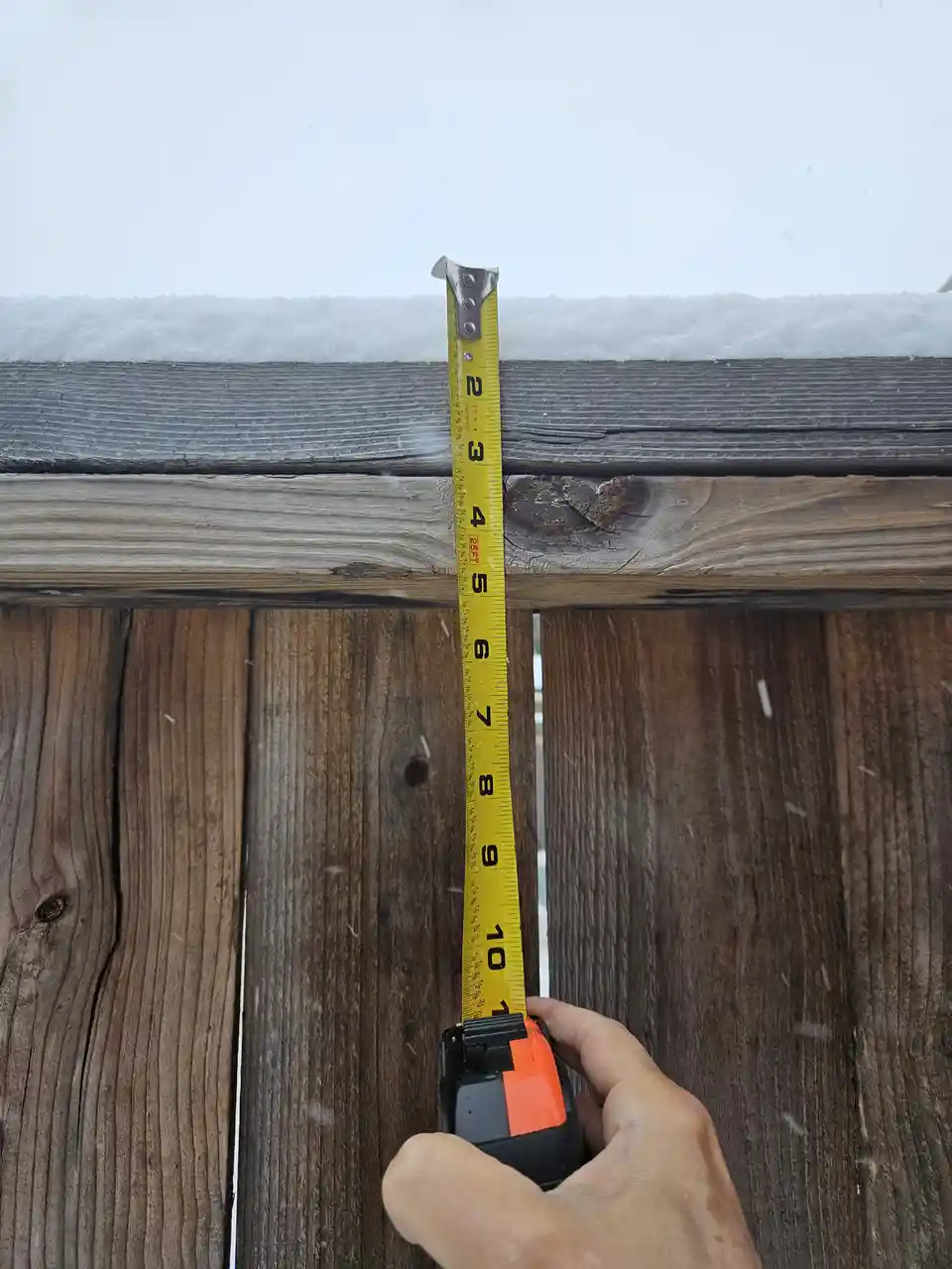 A hand holds a yellow tape measure vertically against a wooden fence, measuring the depth of snow piled on top of the fence…