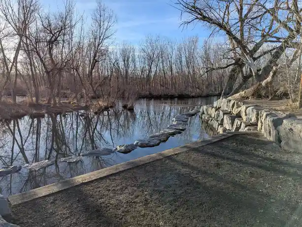 A serene outdoor photo shows a path of stepping stones crossing a calm, reflective pond surrounded by bare winter trees…
