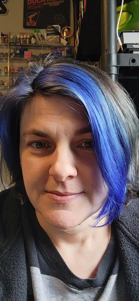 A close-up selfie of a person with short, partially dyed blue hair, looking directly at the camera with a subtle, slightly…
