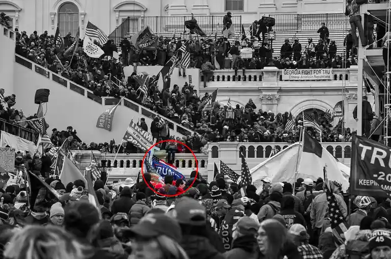 A black-and-white photo of a large crowd of protesters on the steps of the U.S. Capitol, many holding Trump flags and signs…