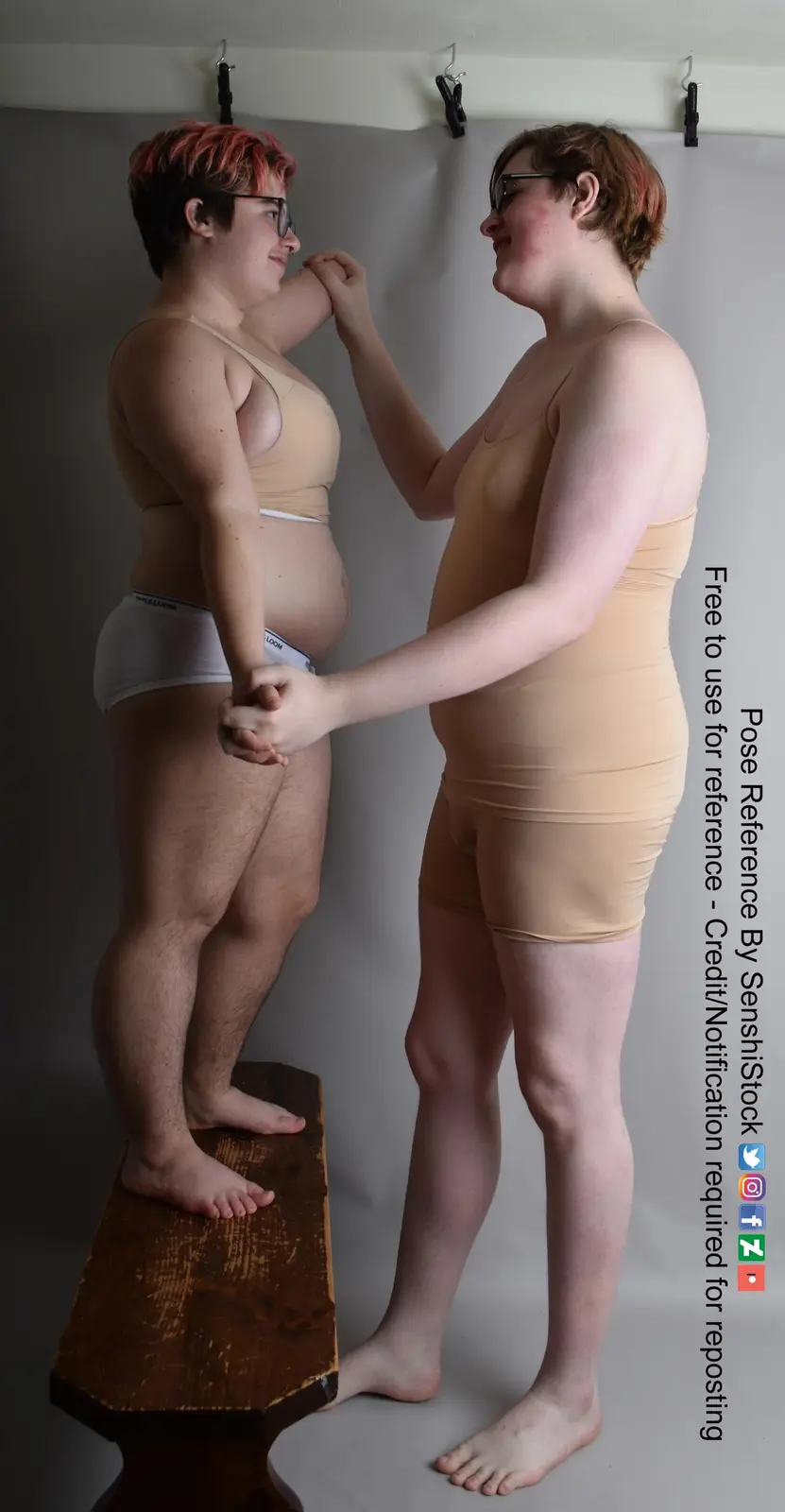 Two people stand facing each other against a gray backdrop; one person stands on a wooden block while the other adjusts…