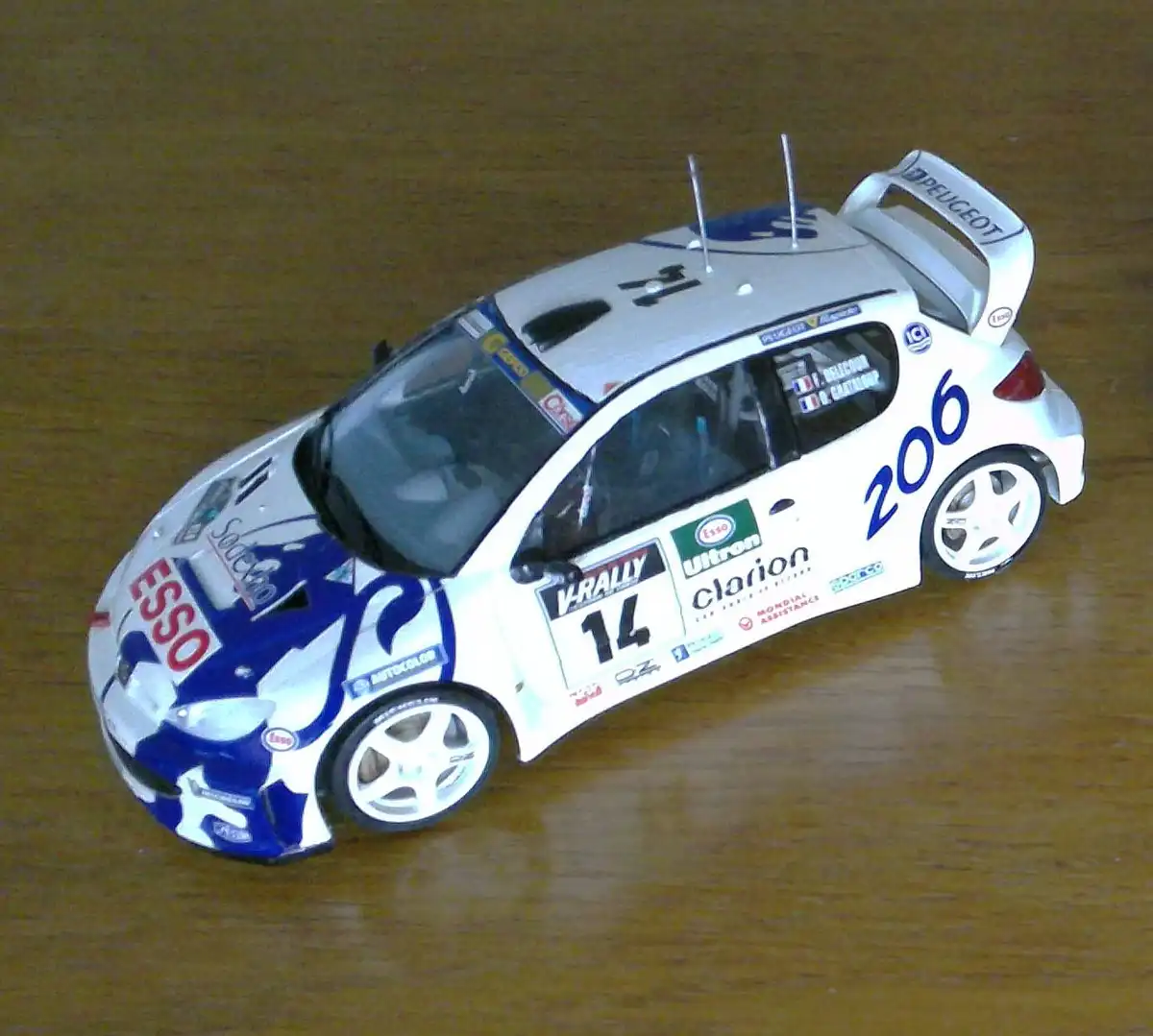 A white and blue scale model of a Peugeot 206 rally car, featuring sponsor logos like ESSO and Clarion, sits on a wooden…