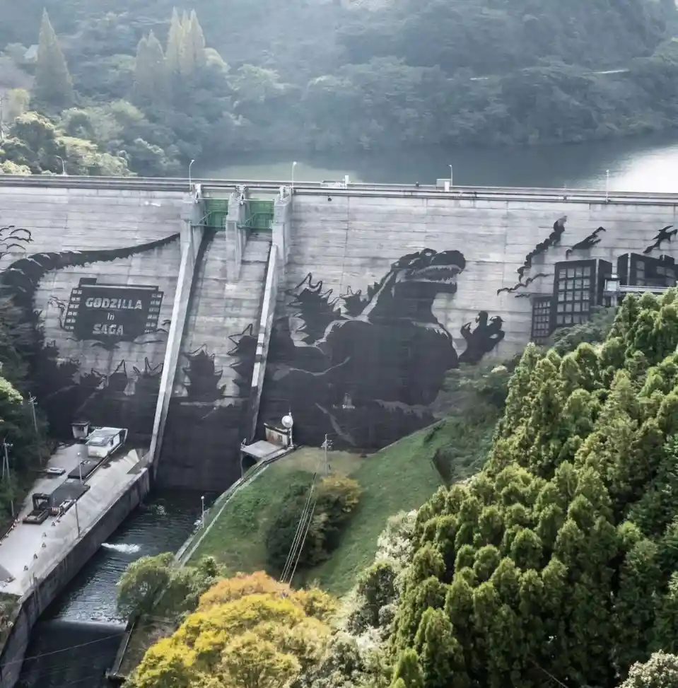 A large concrete dam is painted with a massive black silhouette of Godzilla, appearing to be attacking a city, with the text…