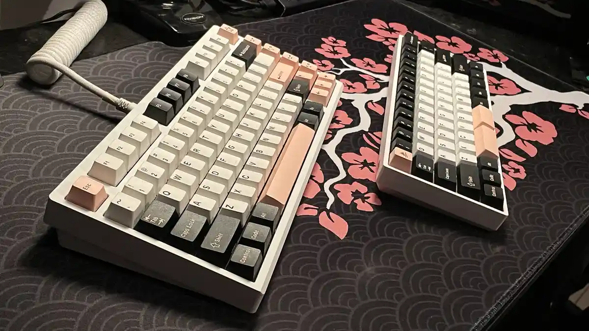 Two mechanical keyboards with mismatched keycaps sit side by side on a dark desk mat with pink cherry blossom patterns. The…