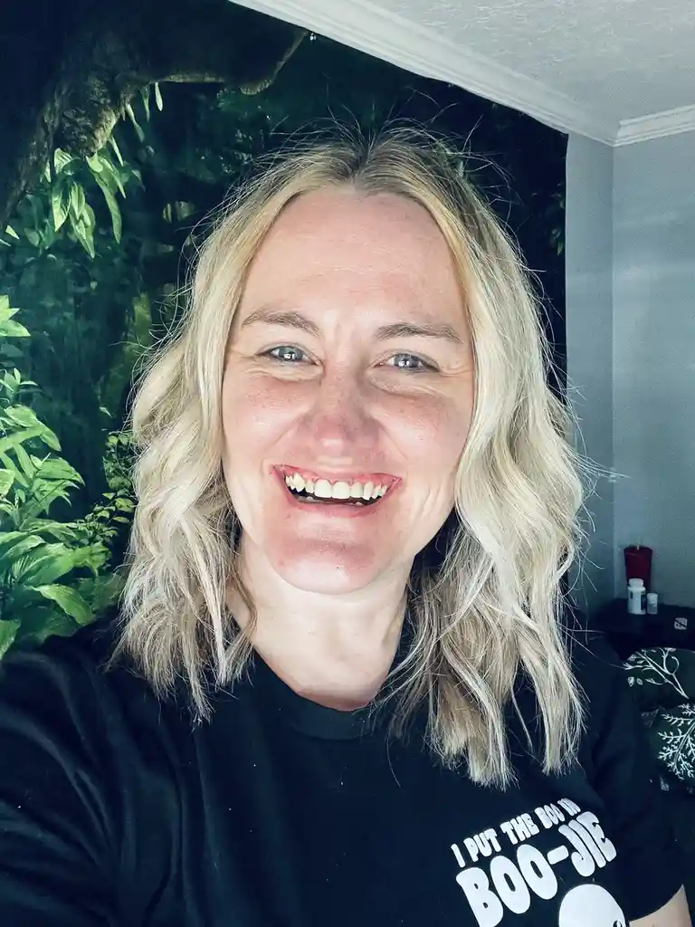 A woman with blonde hair smiles broadly at the camera while wearing a black t-shirt that reads 'I PUT THE BOO IN BOO-JIE'…