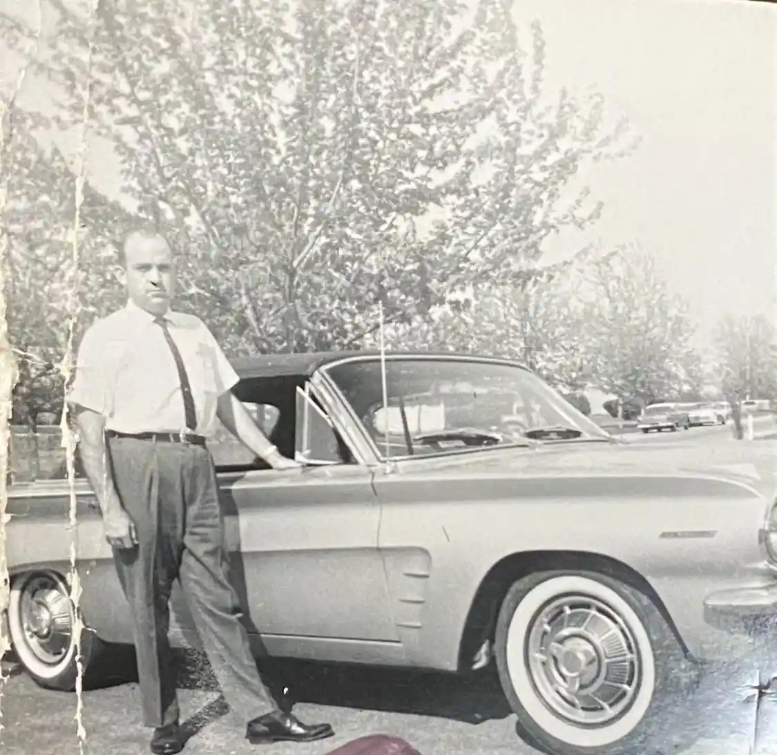 A black-and-white vintage photo shows a man in a short-sleeved shirt and tie standing beside a classic 1960s convertible…