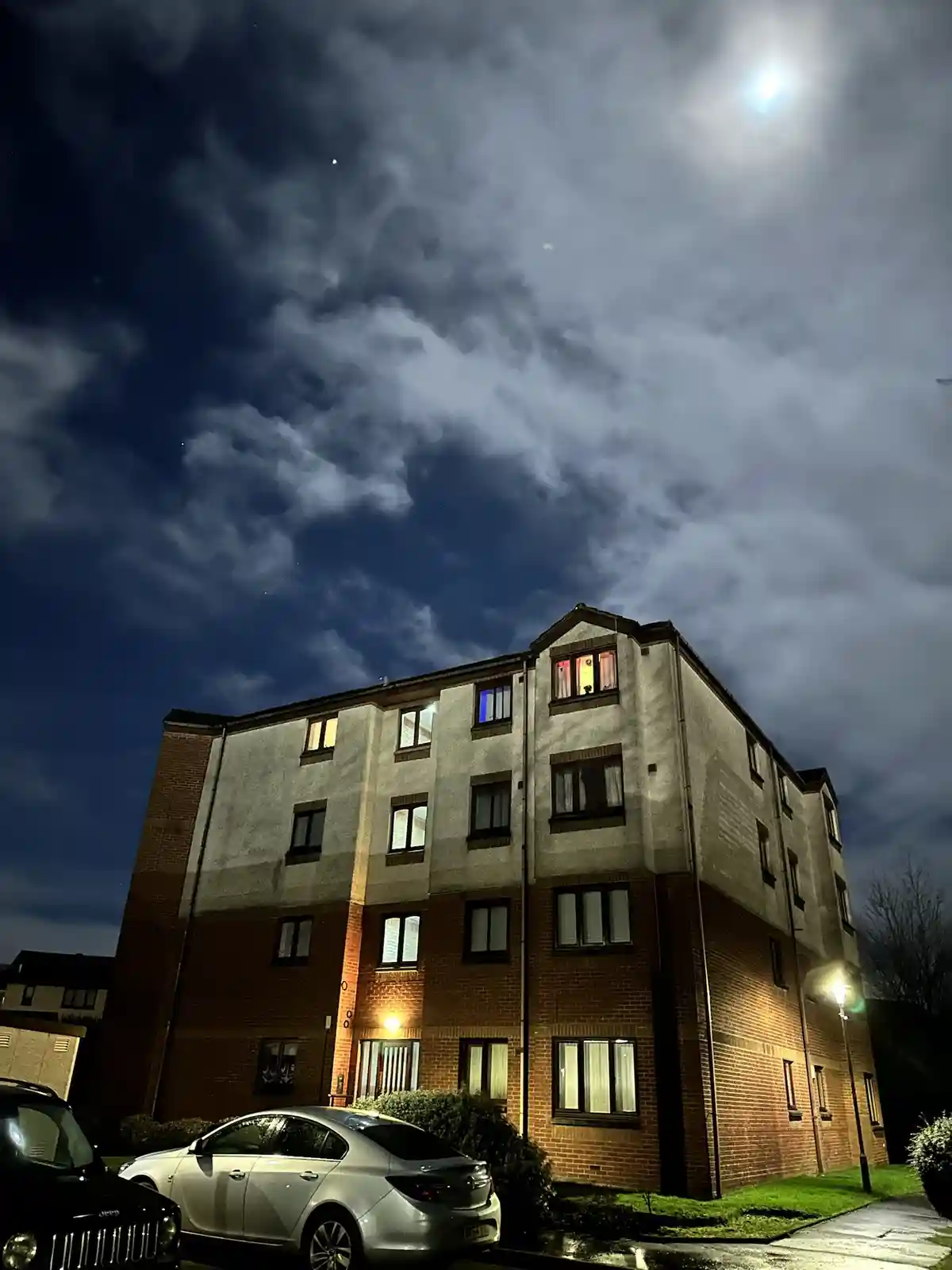 A low-angle night photo shows a multi-story brick apartment building under a cloudy sky with a bright moon visible through…
