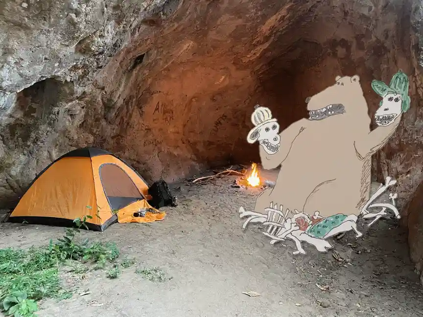 The image shows a cave with an orange tent and a small campfire. Superimposed over the scene is a cartoon bear wearing a…