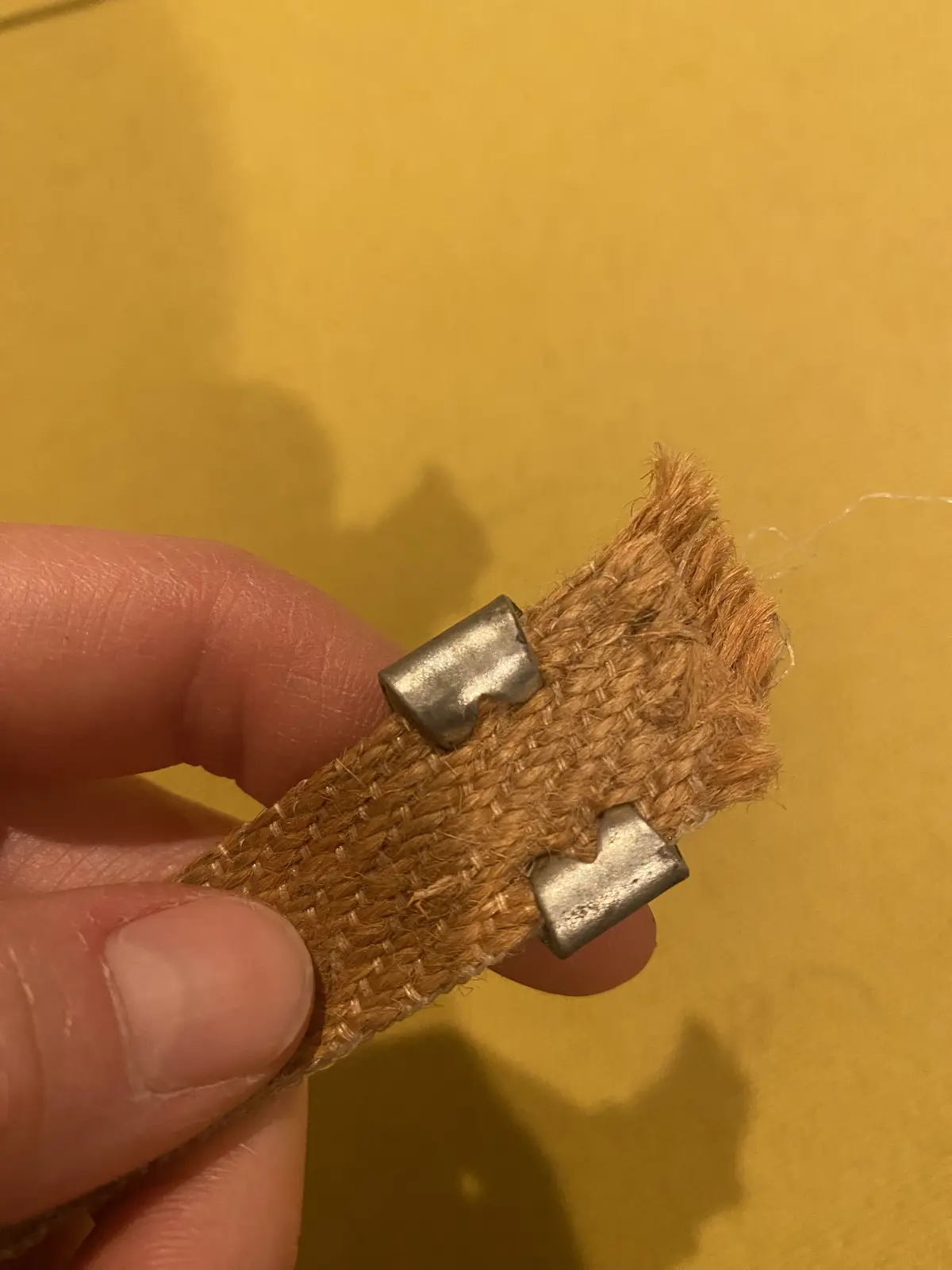 A close-up photo shows a person's fingers holding a frayed, woven brown strap with two metal D-rings attached, against a…