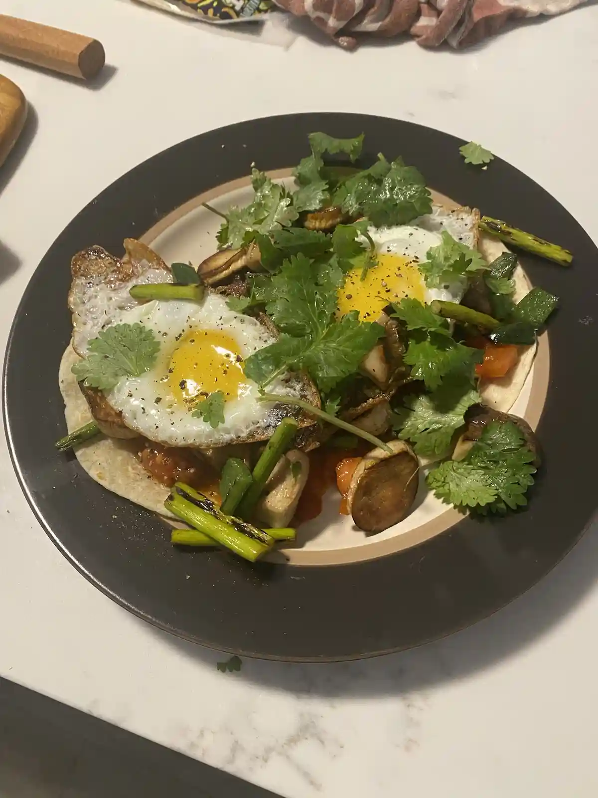 A close-up photo of a plate of food featuring two sunny-side-up eggs, sautéed mushrooms, green onions, and fresh cilantro…