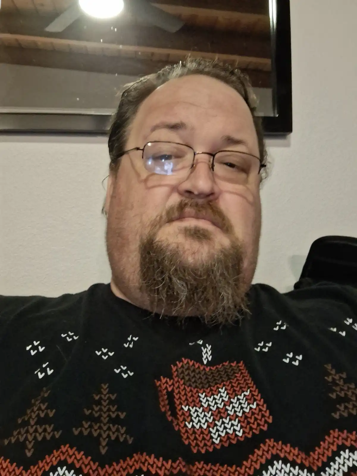 A man with glasses and a beard wearing a black sweater with a red and white patterned design looks directly at the camera…