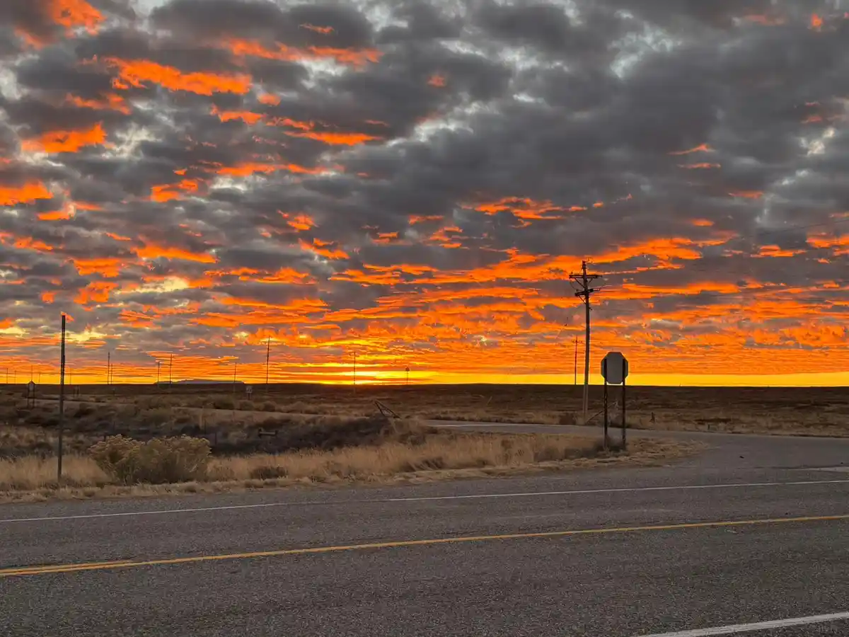 A dramatic sunset with fiery orange and red clouds over a rural road, featuring a stop sign and utility poles in the…