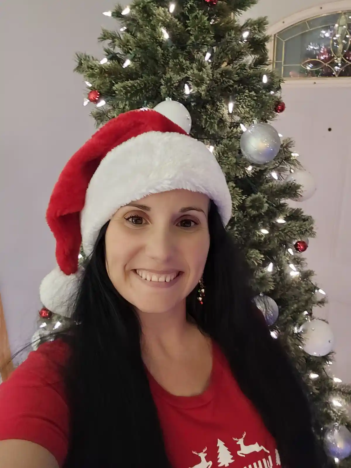 A woman with long dark hair smiles at the camera while wearing a red and white Santa hat and a red t-shirt with white…