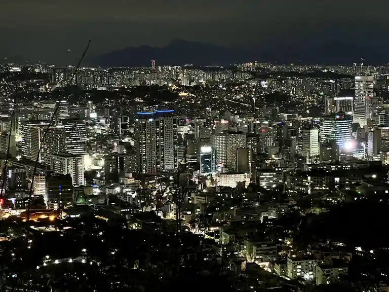 A nighttime cityscape photo showing a sprawling urban area with numerous illuminated buildings and streets, viewed from an…