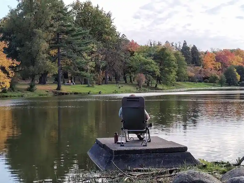 A person sits in a folding chair on a concrete platform at the edge of a calm lake, facing away from the camera toward…