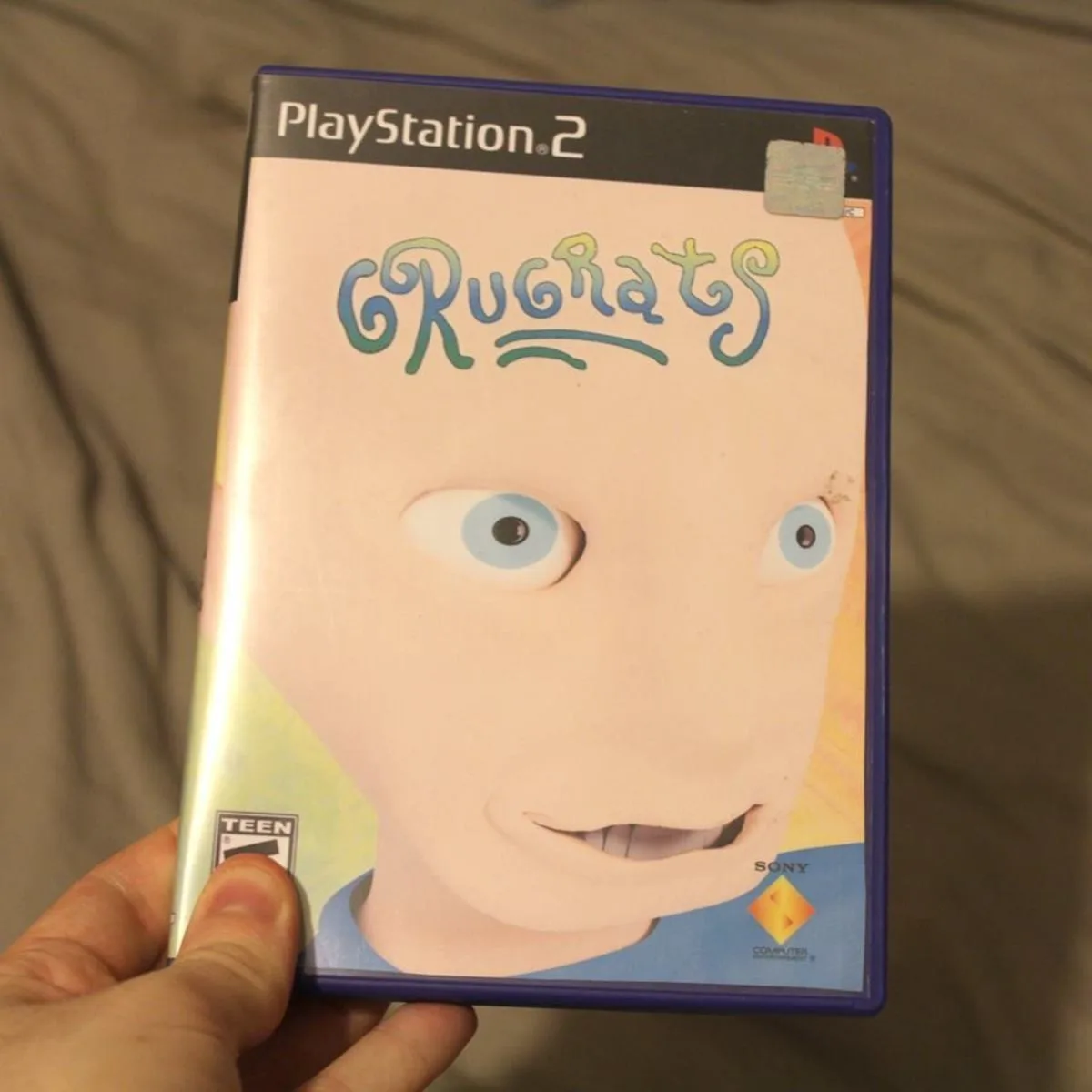 A hand holds a PlayStation 2 game case for 'Rugrats', showing a close-up of the character Tommy Pickles with large blue eyes…