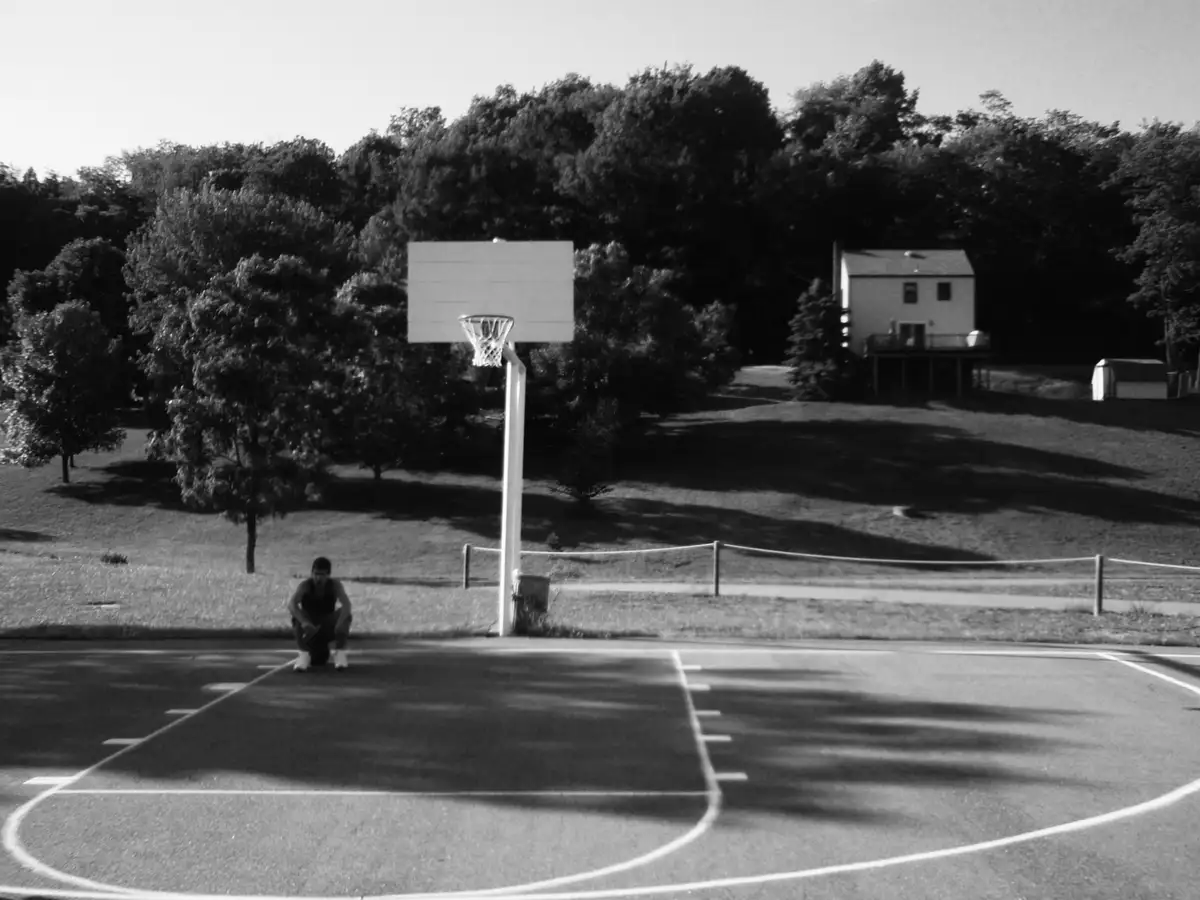 A black-and-white photo shows a person crouching on an outdoor basketball court, holding a basketball, with a hoop in the…