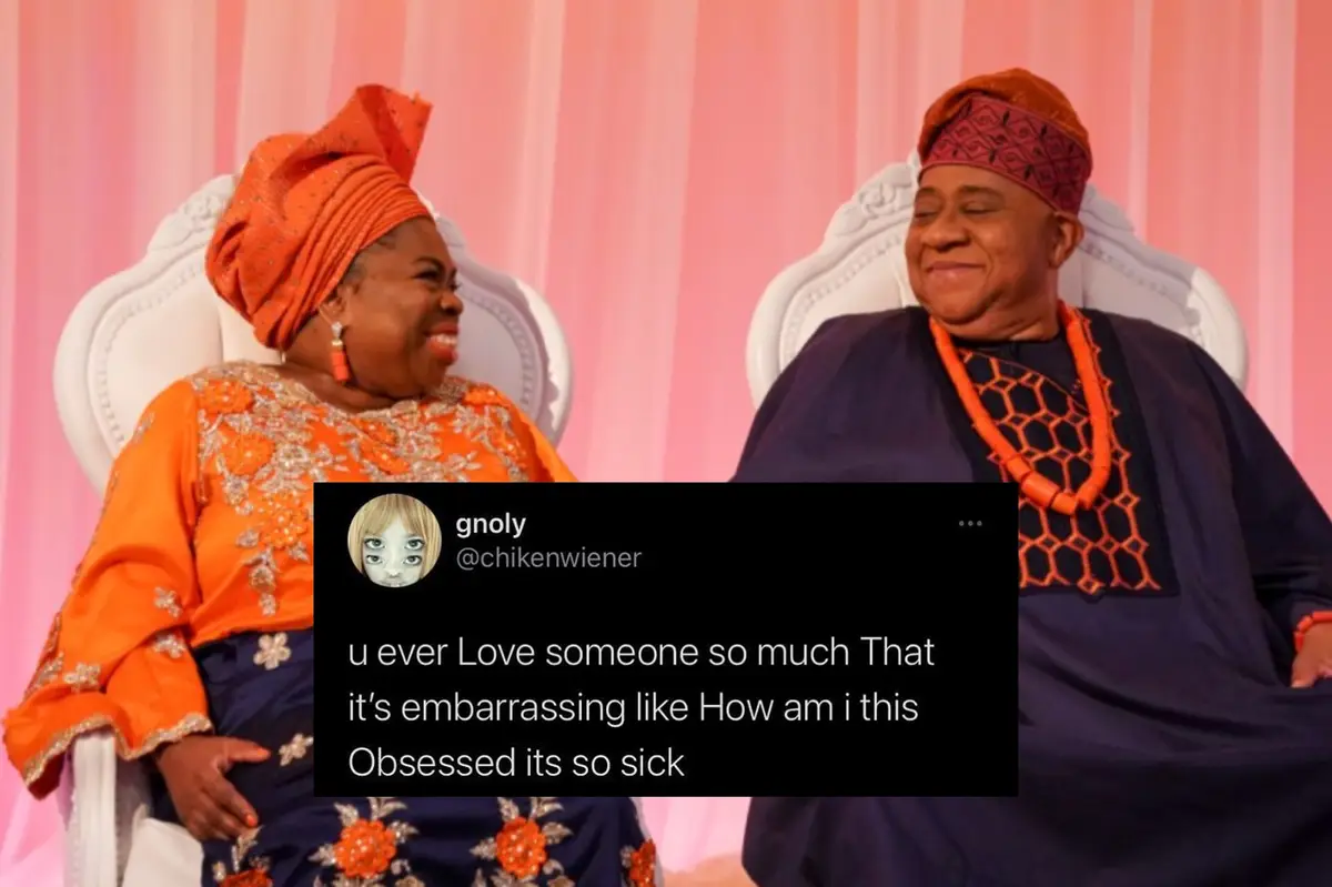 Two people, a woman in orange traditional attire and a man in dark blue with orange beads, sit in ornate white chairs…