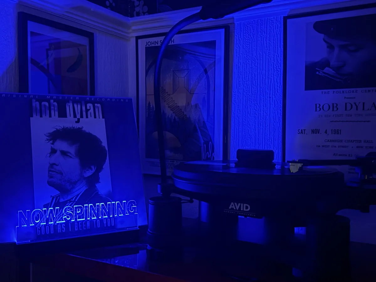 A dimly lit room bathed in blue light features a turntable with a vinyl record, surrounded by framed posters including one…
