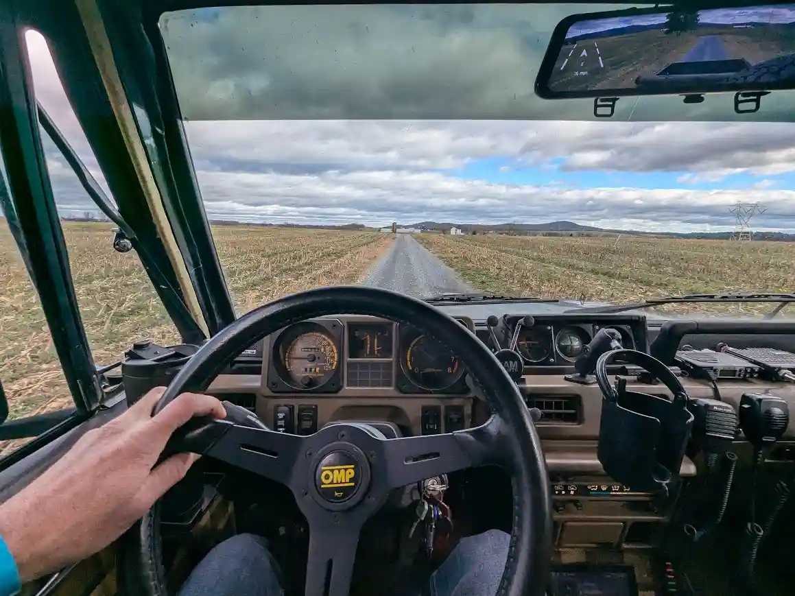 A first-person view from inside a vehicle, likely a tractor or heavy truck, showing a hand on a black OMP steering wheel…