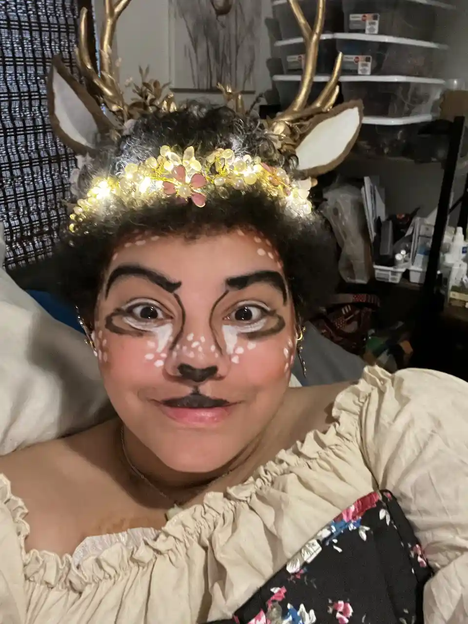 A person with dark curly hair is wearing a golden deer antler headpiece with white ears and a glowing floral crown. Their…