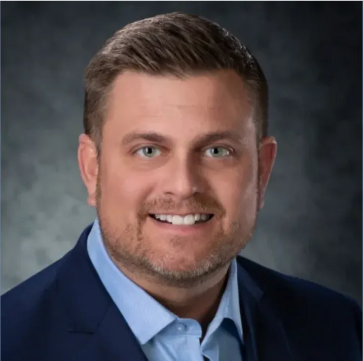 A smiling man with short brown hair and a light beard, wearing a blue suit jacket over a light blue collared shirt, poses…