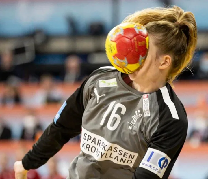 a female handball player wearing a gray jersey with the number 16 and sponsor logos has a red and yellow handball pressed…