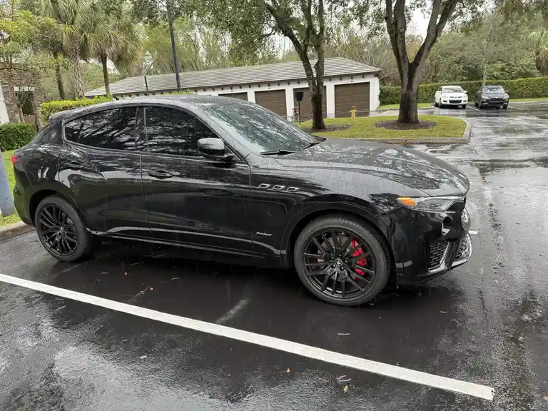 A glossy black Maserati Levante GranSport SUV is parked in a wet parking lot during rain, with water droplets visible on its…