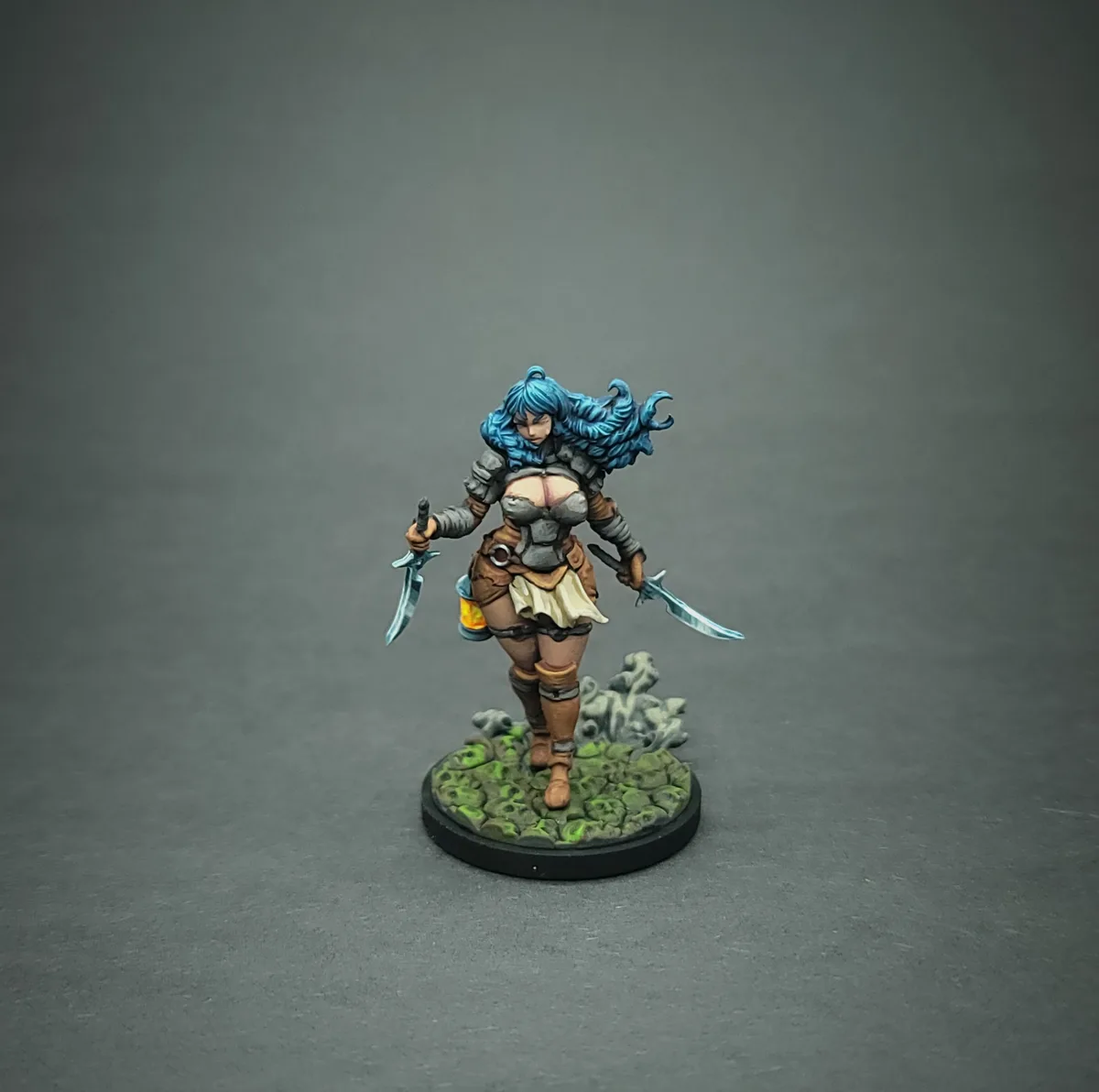 A painted miniature figurine of a female warrior with long blue hair, wearing armor and holding two swords, stands on a…
