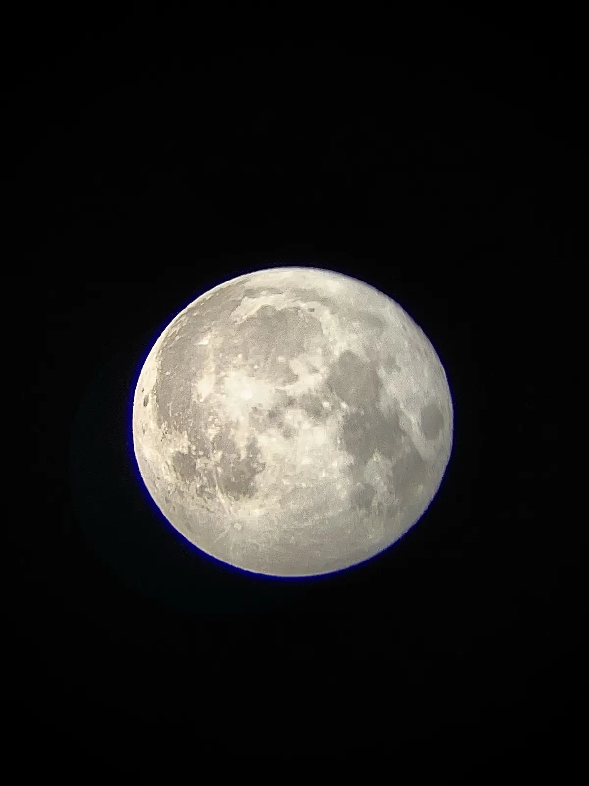 A close-up photograph of the full moon against a black sky, with a subtle blue halo around its edge. The moon's surface…
