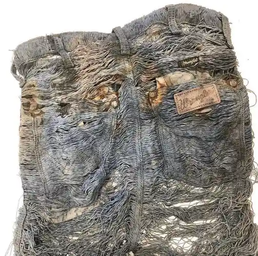 A close-up of a pair of extremely frayed, worn-out Wrangler jeans, showing shredded fabric, exposed stitching, and faded…