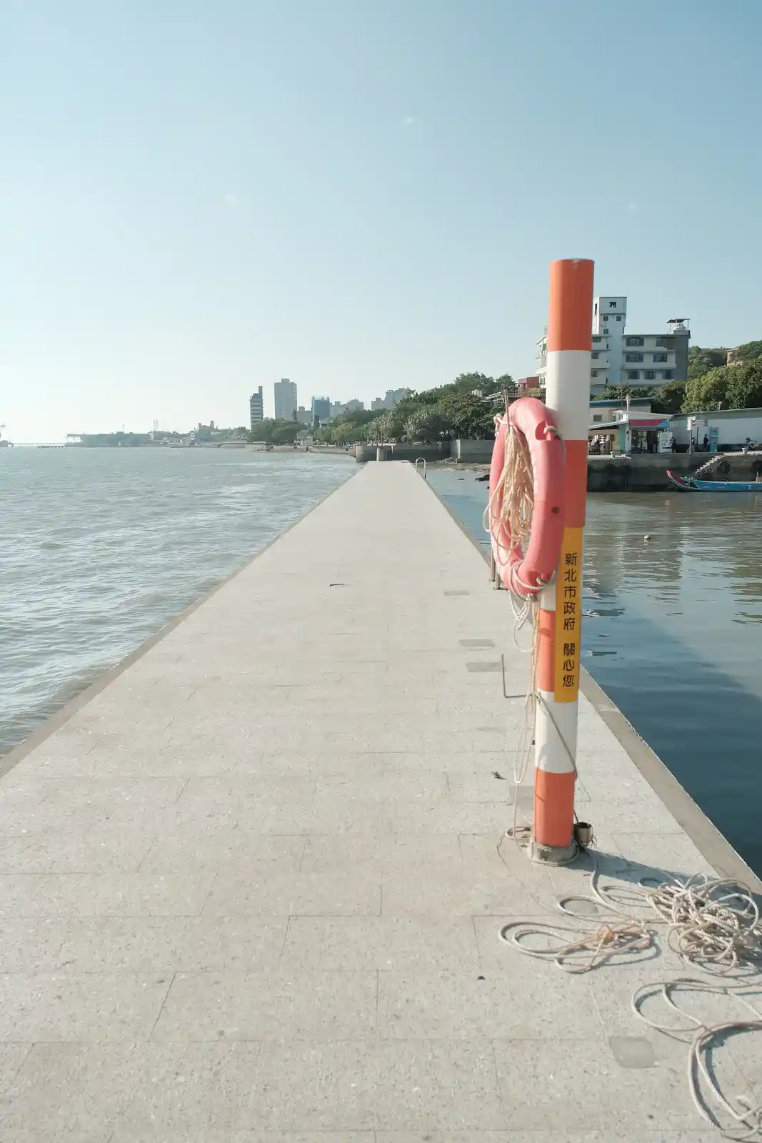 A photo of a concrete pier extending into water under a clear sky, with a red and white striped pole holding a pink…