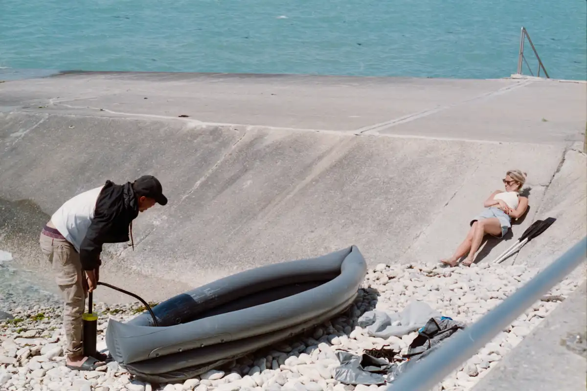 A man on a rocky beach is vigorously pumping air into an inflatable boat while a woman lounges nearby, seemingly unbothered…