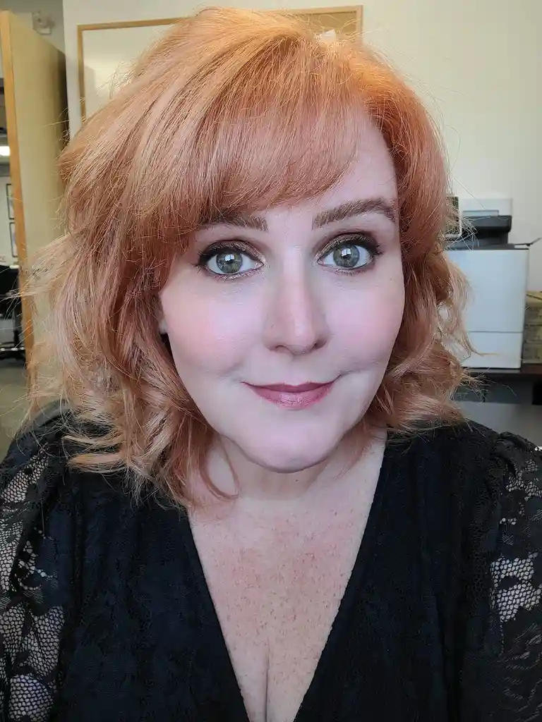 A woman with shoulder-length, wavy copper-red hair and light green eyes looks directly at the camera with a subtle smile…