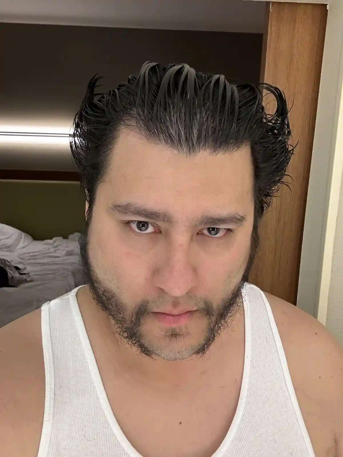 A man with dark, wet-looking hair styled in a quiff and a full beard stares intensely at the camera while wearing a white…
