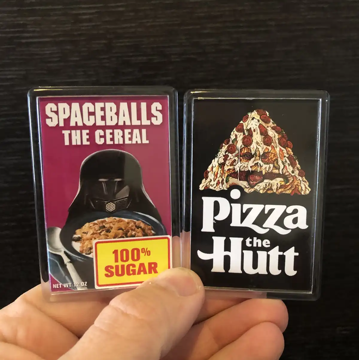 A hand holds two rectangular fridge magnets side-by-side. The left magnet features a parody cereal box design with Darth…