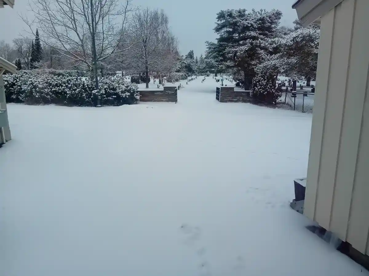 A snowy outdoor scene viewed from a doorway, showing a snow-covered cemetery with tombstones, bare trees, and bushes in the…