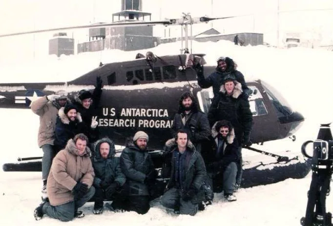 A group of people in heavy winter gear pose for a photo in front of a snow-covered helicopter marked 'U.S. ANTARCTICA…
